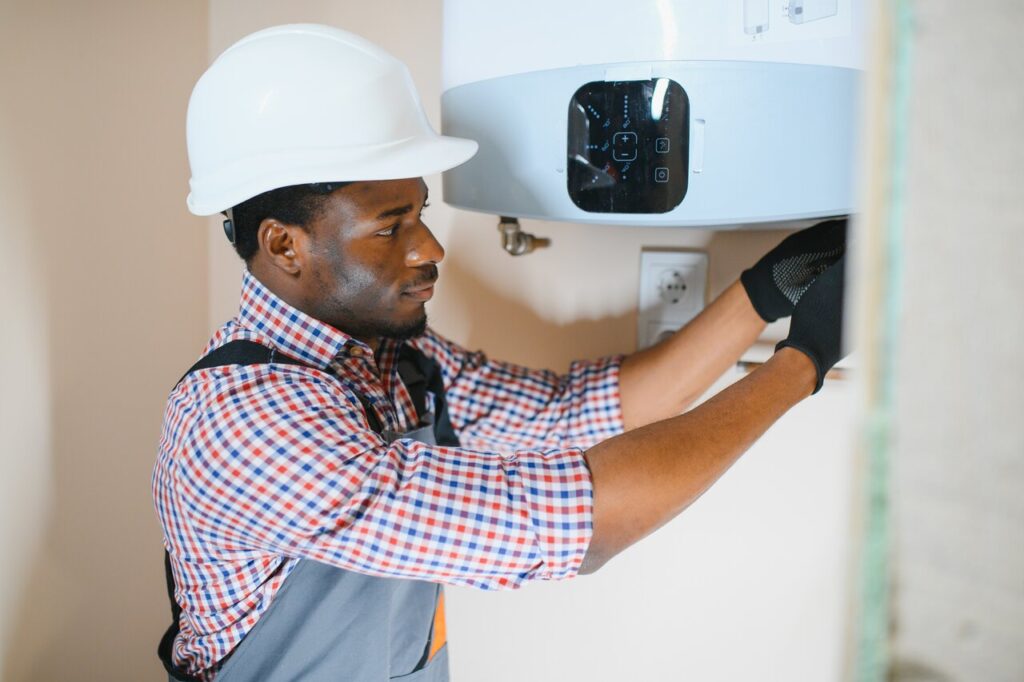 A man wearing a hard hat and overalls repairs a water heater, demonstrating skilled maintenance work.