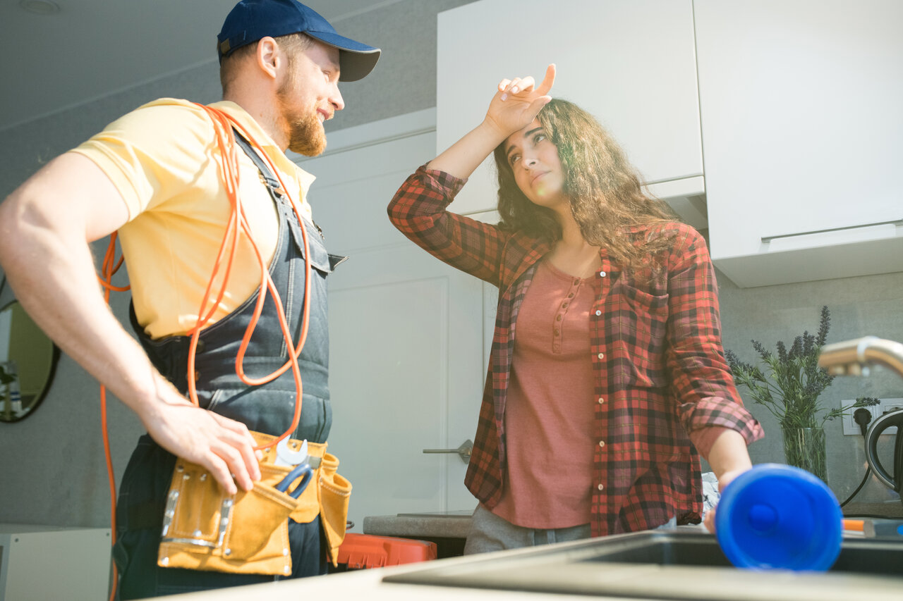 A man and woman stand together in a kitchen, positioned near a sink, engaged in conversation or cooking.